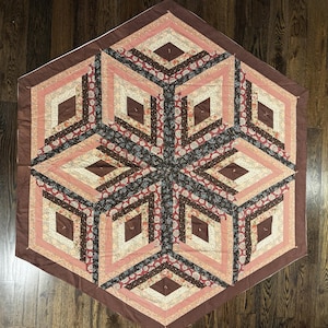 May include: A hexagonal quilt featuring a geometric pattern of diamonds and triangles. The fabric pieces are arranged in shades of brown, cream, and dusty pink, with floral and dotted prints creating a three-dimensional cube effect.