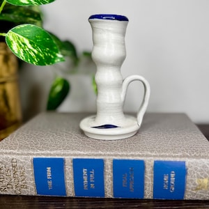 May include: A handmade ceramic candlestick holder. The white candlestick has a blue rim and base, and a small handle. The candlestick is sitting on a book. Green leafy plant in the background.