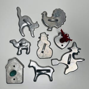 May include: A collection of eight silver-toned metal cookie cutters in various shapes including a cat, turkey, camel, snowman, heart with a red ribbon, witch on a broomstick, house with a green accent, and a horse, each featuring a central handle.
