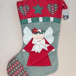 May include: A Christmas stocking with a red top featuring hearts and a green star. The body is green and white checkered fabric with an angel applique. A silver bell hangs from the top.