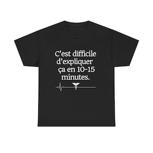 May include: Black t-shirt with white text that reads "C'est difficile d'expliquer ça en 10-15 minutes." A white medical symbol and a heartbeat line are below the text. The shirt is a crew neck.