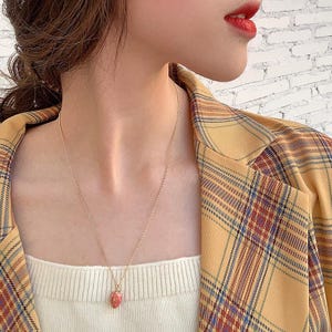 May include: A delicate gold necklace with a small, pink strawberry pendant. The necklace is worn over a white top and against a yellow plaid blazer. The image is a close-up, focusing on the jewelry.