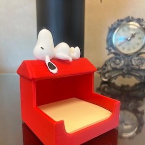 May include: A red, 3D-printed Snoopy doghouse-shaped desk organizer. A white Snoopy figure is lounging on the roof. The organizer has a beige notepad inside. A decorative clock and a black vase are in the background.