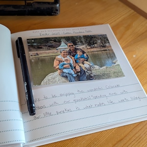 May include: An open journal with a photograph and handwritten text. A black pen rests on the left page. The photo shows a family by a lake. Text includes "Easter 2025-Green Monde" and handwritten notes about enjoying Colorado.