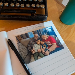 May include: A black Sharpie pen rests on an open photo album with a family photo. The album is open to a page with lined paper and a printed date and location. An antique typewriter is in the background.