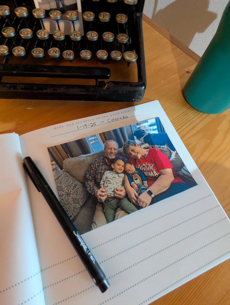 May include: A black Sharpie pen rests on an open photo album with a family photo. The album is open to a page with lined paper and a printed date and location. An antique typewriter is in the background.