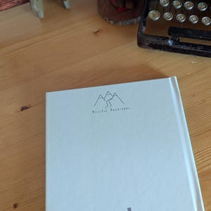 May include: A white book with the text "Blissful Backroads" and a mountain illustration. The book is on a wooden surface, with a vintage typewriter and a wooden bear figurine in the background.
