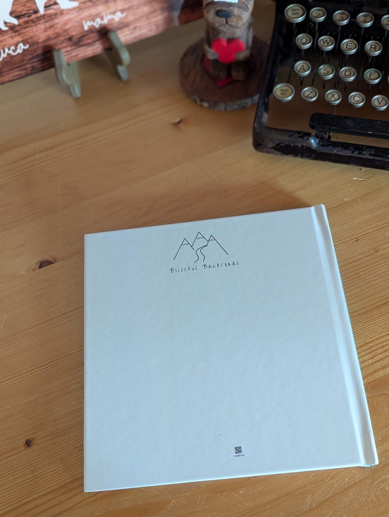 May include: A white book with the text "Blissful Backroads" and a mountain illustration. The book is on a wooden surface, with a vintage typewriter and a wooden bear figurine in the background.