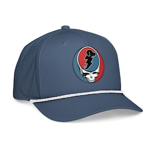 May include: A blue baseball cap with a white rope detail and a curved brim. The cap features a circular emblem with a skull and lightning bolt design in red, blue, and white.