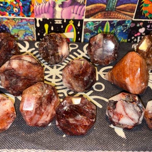 May include: A collection of eleven polished, faceted stones featuring deep red, brown, and white marbled patterns. Each stone displays a unique crystalline structure with smooth, reflective surfaces and varied geometric shapes arranged on a dark fabric.