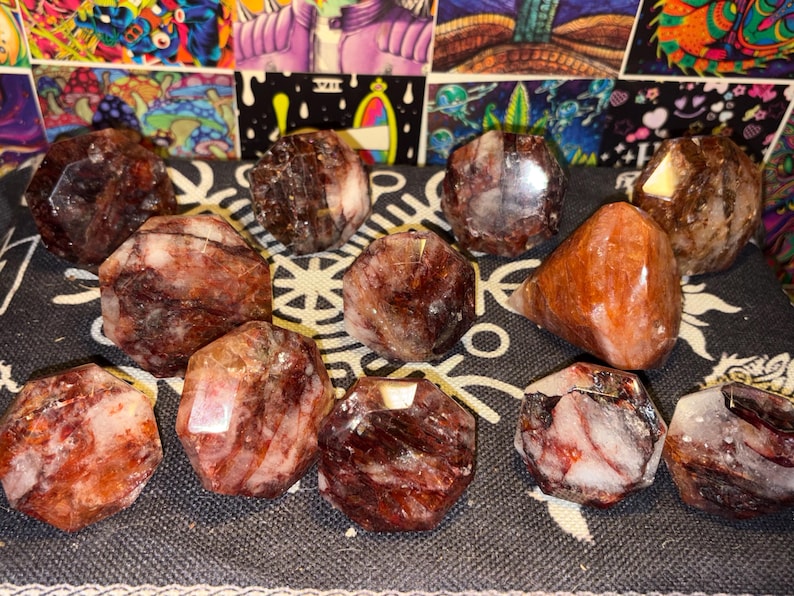 May include: A collection of eleven polished, faceted stones featuring deep red, brown, and white marbled patterns. Each stone displays a unique crystalline structure with smooth, reflective surfaces and varied geometric shapes arranged on a dark fabric.
