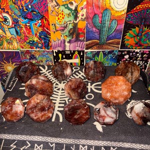 May include: A collection of twelve faceted, polished stones arranged on a dark fabric surface. The stones feature a mix of deep reddish-brown, translucent white, and bright orange hues, each cut into a multi-sided, geometric shape with flat surfaces.