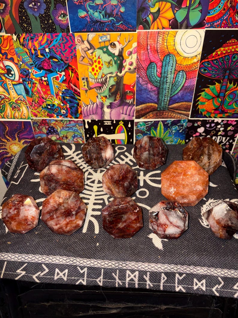 May include: A collection of twelve faceted, polished stones arranged on a dark fabric surface. The stones feature a mix of deep reddish-brown, translucent white, and bright orange hues, each cut into a multi-sided, geometric shape with flat surfaces.