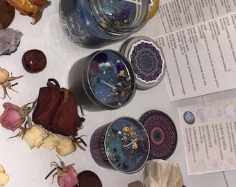 May include: Several candles in glass jars and tins with decorative lids. The candles are blue with decorative elements. Dried flowers and crystals surround the candles, along with a paper with text.