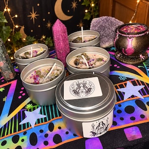 May include: Four silver metal candle tins filled with wax, herbs, and crystals, accompanied by a small black cauldron, a tall pink crystal point, a dark green crystal point, and a large amethyst cluster. One tin features a circular label with a goddess illustration.