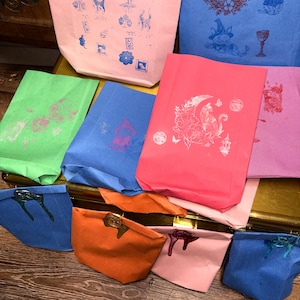 May include: A collection of colorful paper bags, each adorned with unique stamped designs in various colors. The bags are sealed with wax and arranged on a wooden surface. The designs include cats, moons, and other whimsical illustrations.