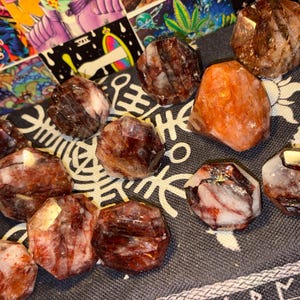 May include: A collection of faceted, polished stones featuring deep red, brown, and white marbled patterns. These stones have a geometric, multi-sided shape with smooth surfaces, displaying natural mineral inclusions and varied color gradients throughout.