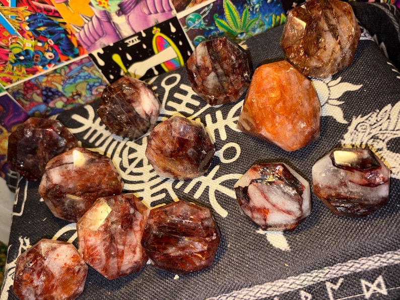 May include: A collection of faceted, polished stones featuring deep red, brown, and white marbled patterns. These stones have a geometric, multi-sided shape with smooth surfaces, displaying natural mineral inclusions and varied color gradients throughout.