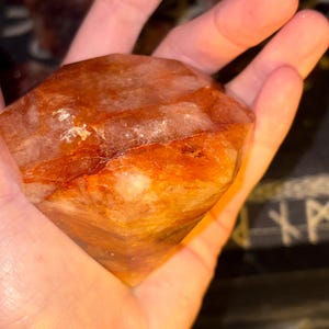 May include: A faceted, diamond-shaped crystal with a vibrant orange and reddish-brown hue. The stone features internal fractures and natural inclusions, displaying a semi-translucent appearance with varying shades of amber and deep rust throughout.