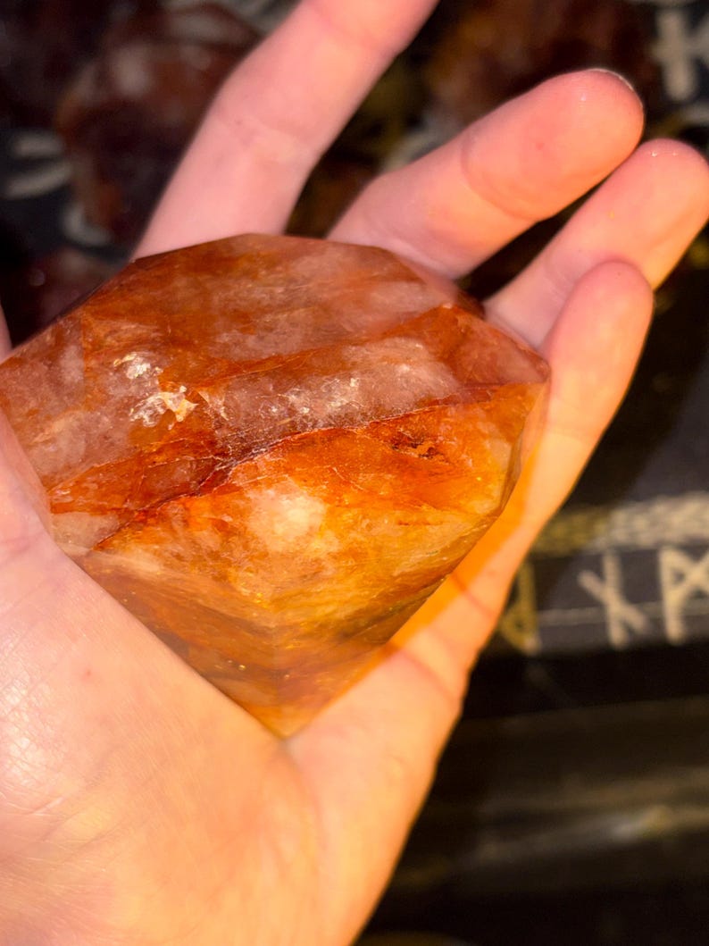 May include: A faceted, diamond-shaped crystal with a vibrant orange and reddish-brown hue. The stone features internal fractures and natural inclusions, displaying a semi-translucent appearance with varying shades of amber and deep rust throughout.
