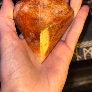 May include: A faceted, diamond-shaped gemstone with a warm, translucent orange and amber hue. The stone features internal fractures and inclusions, creating a complex, multi-layered appearance with varying shades of deep gold and burnt orange.