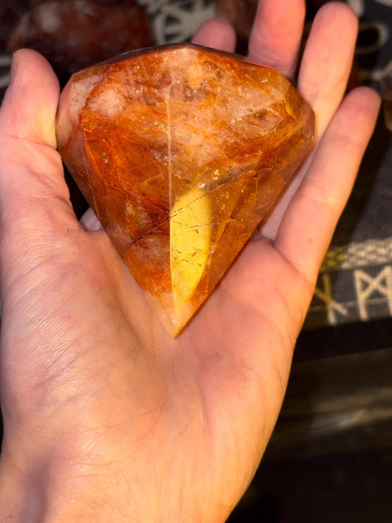 May include: A faceted, diamond-shaped gemstone with a warm, translucent orange and amber hue. The stone features internal fractures and inclusions, creating a complex, multi-layered appearance with varying shades of deep gold and burnt orange.