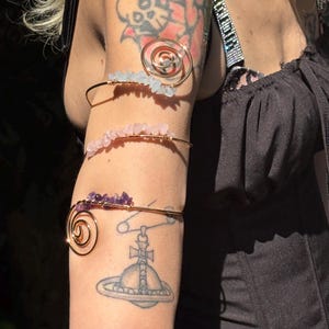 May include: Three gold-toned arm cuffs adorned with gemstones. The top cuff features a spiral design and light blue stones. The middle cuff has pink stones, and the bottom cuff has purple stones. The arm cuffs are worn on the upper arm.