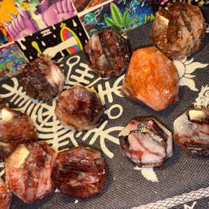 May include: A collection of faceted, polished stones in shades of deep red, brown, and translucent white, with one prominent orange-hued stone. These stones feature irregular, multi-sided geometric shapes with visible internal mineral inclusions.