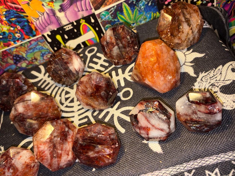 May include: A collection of faceted, polished stones in shades of deep red, brown, and translucent white, with one prominent orange-hued stone. These stones feature irregular, multi-sided geometric shapes with visible internal mineral inclusions.