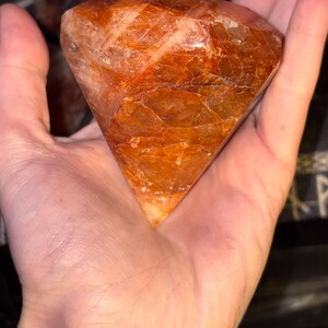 May include: A faceted, diamond-shaped gemstone with a deep burnt orange and reddish-brown hue. The stone features a polished surface with visible internal fractures and natural crystalline inclusions, displaying a translucent, warm color palette.