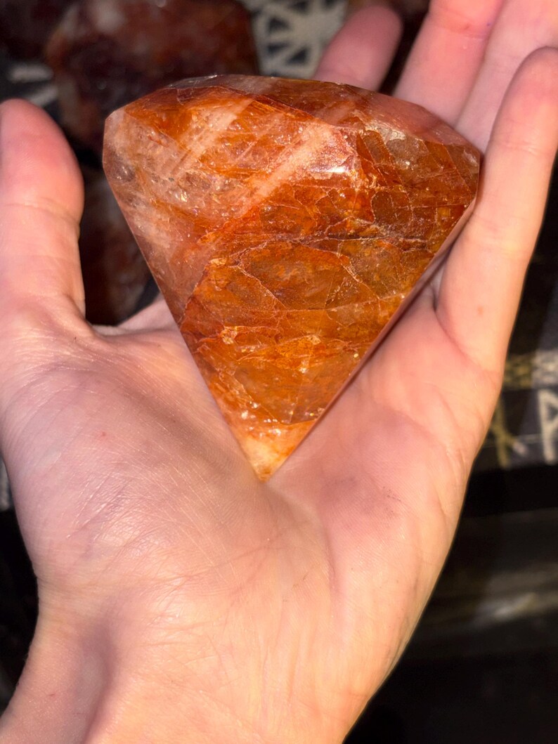 May include: A faceted, diamond-shaped gemstone with a deep burnt orange and reddish-brown hue. The stone features a polished surface with visible internal fractures and natural crystalline inclusions, displaying a translucent, warm color palette.
