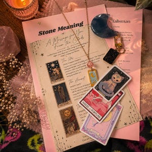 May include: A gold-tone necklace with a pink bead and a rectangular pendant featuring a moon and star design, resting on a printed card titled Stone Meaning and A Message for You From the Cards, alongside three tarot cards and a blue crescent moon.