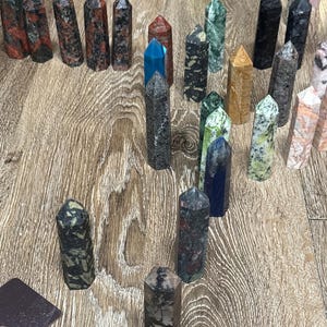 May include: A collection of various gemstone obelisks in shades of red, black, green, blue, and tan. The obelisks are arranged on a wooden surface, showcasing their unique patterns and textures. These decorative crystals are ideal for home decor or spiritual practices.