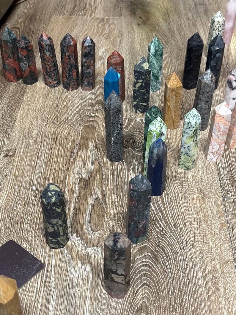 May include: A collection of various gemstone obelisks in shades of red, black, green, blue, and tan. The obelisks are arranged on a wooden surface, showcasing their unique patterns and textures. These decorative crystals are ideal for home decor or spiritual practices.