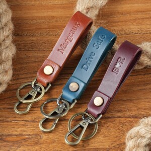 May include: Three leather keychains in brown, blue, and burgundy. Each keychain has a metal clasp and a leather strap with text: "Margeaux", "Drive Safe", and "BVR". The keychains are on a wooden surface.