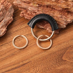 May include: A black leather key ring with gold-colored metal rings. The key ring has a curved leather handle with stitched detailing. Two additional gold-colored rings are attached.