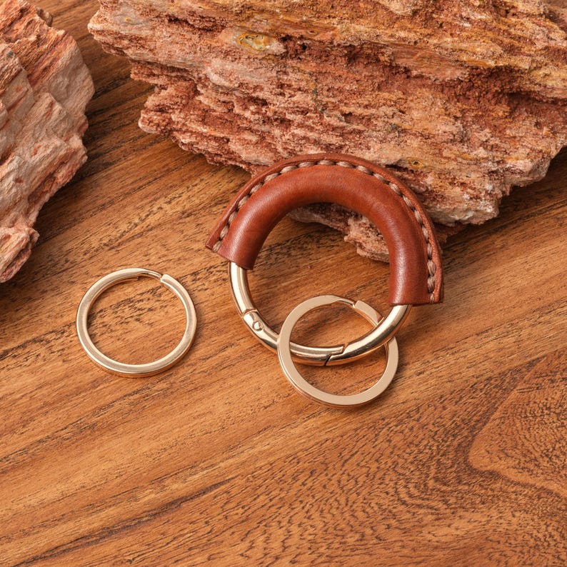 May include: A brown leather key ring with a gold-colored metal clasp and two gold-colored key rings. The leather is stitched with a lighter brown thread. The key ring is on a wooden surface.