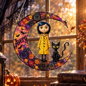 May include: A crescent moon-shaped hanging decoration featuring a mosaic of colorful buttons and small bat silhouettes. The name Avery is printed on the left side. A figure with blue hair in a yellow raincoat and a black cat stand on the moon.