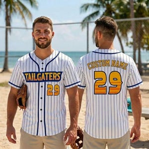 May include: White baseball jersey with blue pinstripes, featuring the text "TAILGATERS" and the number "29" in orange and yellow. The jersey has a button-up front and blue trim. A baseball glove is visible.