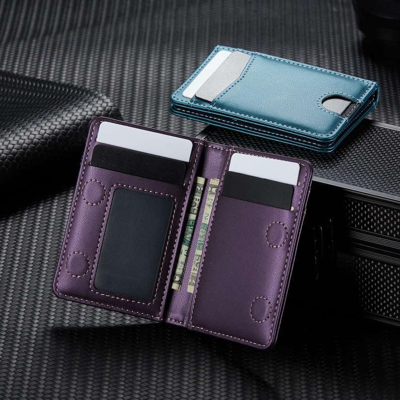 May include: Purple bi-fold wallet with multiple card slots and a clear ID window. The wallet is open, revealing several cards and cash. A teal cardholder is visible in the background. The wallets are made of leather and have a stitched design.