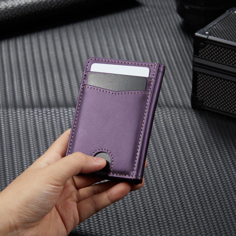 May include: A purple leather cardholder with a minimalist design. The wallet features a slot for cards and a push-button mechanism. The stitching is visible around the edges. The wallet is designed to hold cards.
