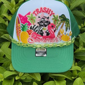 May include: A green and white trucker hat with a colorful summer-themed design. The hat features a raccoon, flamingo, and tropical elements. The text "TRASHY Summer" is displayed across the front. The hat has a curved brim and a mesh back.