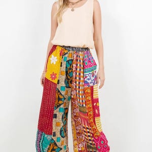 Op de afbeelding: Een vrouw draagt een crèmekleurige tanktop en een patchwork palazzo broek. De broek met wijde pijpen heeft een verscheidenheid aan kleurrijke stoffen patches in rood, geel, blauw en roze. De broek heeft een elastische tailleband.