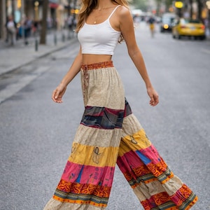 Puede incluir: Una mujer con un top corto blanco y pantalones palazzo estampados de colores. Los pantalones tienen rayas horizontales en beige, amarillo, rosa y rojo con diseños decorativos. Lleva sandalias marrones.