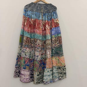 May include: A long, tiered skirt featuring a patchwork design with various colourful patterns, including paisley, floral, and geometric prints in shades of blue, red, green, and purple. The skirt has an elasticated waistband with a drawstring tie.