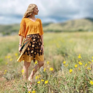 May include: An orange V-neck top and a wrap skirt with a brown and gold leaf pattern. The skirt has a yellow border. The outfit is worn outdoors in a field of yellow flowers.
