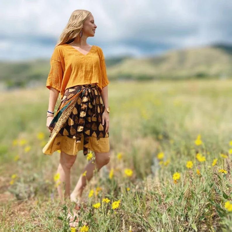 May include: An orange V-neck top and a wrap skirt with a brown and gold leaf pattern. The skirt has a yellow border. The outfit is worn outdoors in a field of yellow flowers.