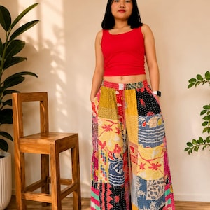 Könnte beinhalten: Eine Frau trägt ein rotes Tanktop und eine Patchwork-Palazzohose. Die weite Hose hat eine Vielzahl von bunten Stoffflicken mit floralen und geometrischen Mustern. Ein Holzstuhl und Topfpflanzen sind im Hintergrund.