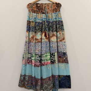 May include: A long, tiered maxi skirt featuring a patchwork design with various colourful patterns, including paisley, floral, and geometric prints in shades of blue, brown, green, and cream. The skirt has an elasticated, smocked waistband with a drawstring.