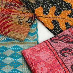 May include: A collection of colorful, patterned textiles. The fabrics feature intricate stitching and designs in shades of red, orange, blue, and black. The textiles appear to be folded or layered, showcasing the variety of patterns.